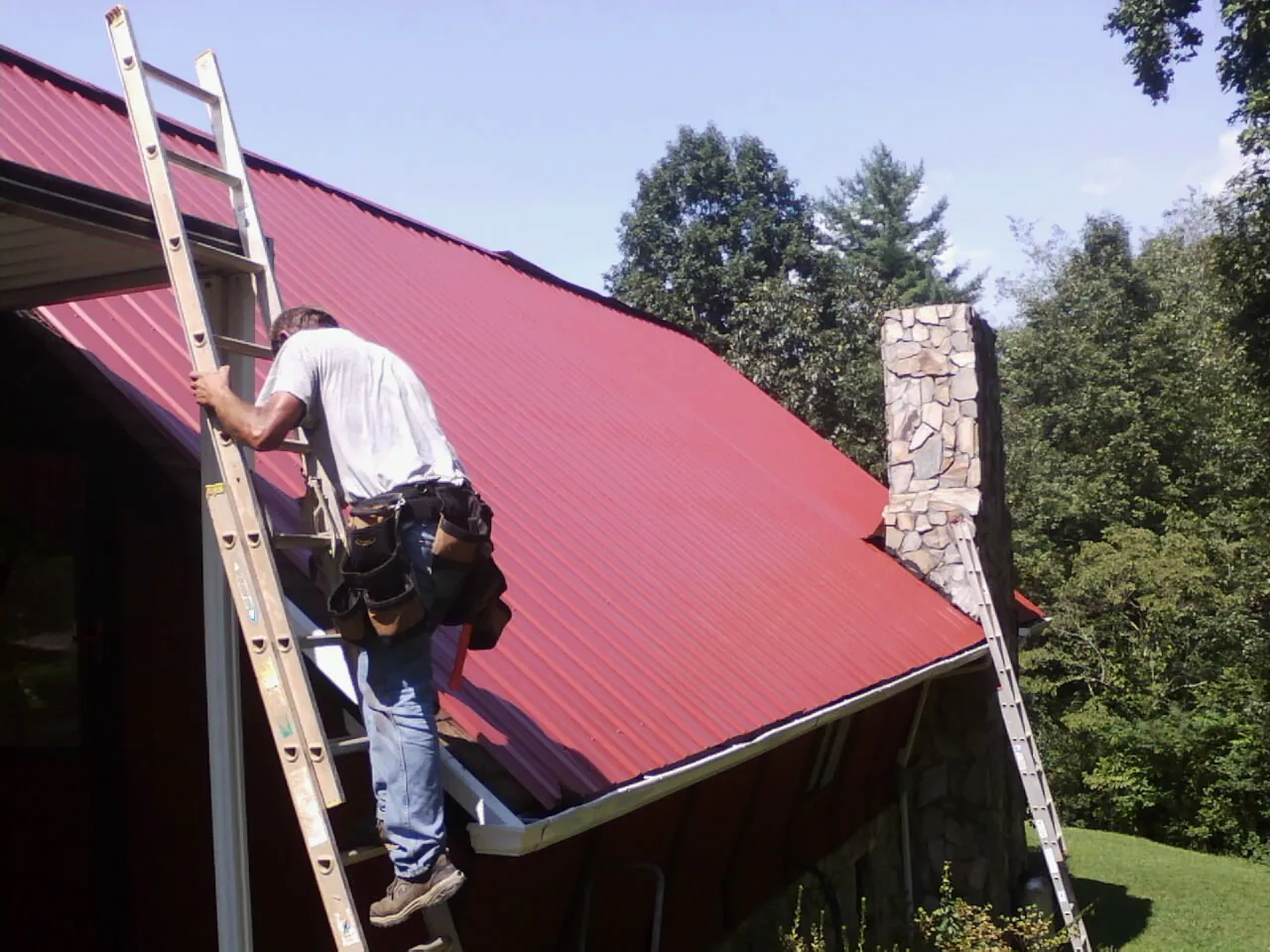Roofer on a ladder beside a red metal roof and stone chimney — Soffit & Fascia Repair by Roofing Brothers in Maceo